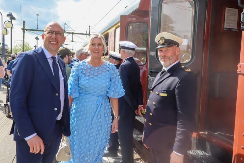 Tre personer står i närheten av ett tåg. En man i blå kostym och glasögon ler. En kvinna i blå klänning ler. En man i marinuniform med vit hatt står till höger.