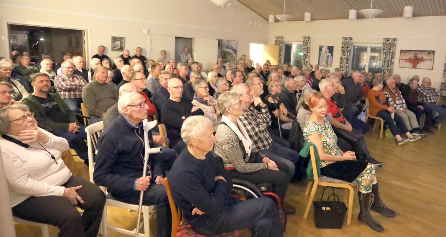 En stor grupp äldre vuxna sitter tätt tillsammans på stolar i ett ljust rum och tittar uppmärksamt på något utanför bild.