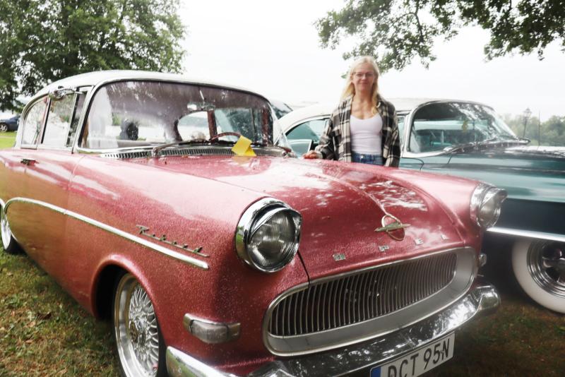 En person står bredvid en röd Opel Rekord från förr som står parkerad på gräset, med en annan klassisk bil som syns delvis i bakgrunden.