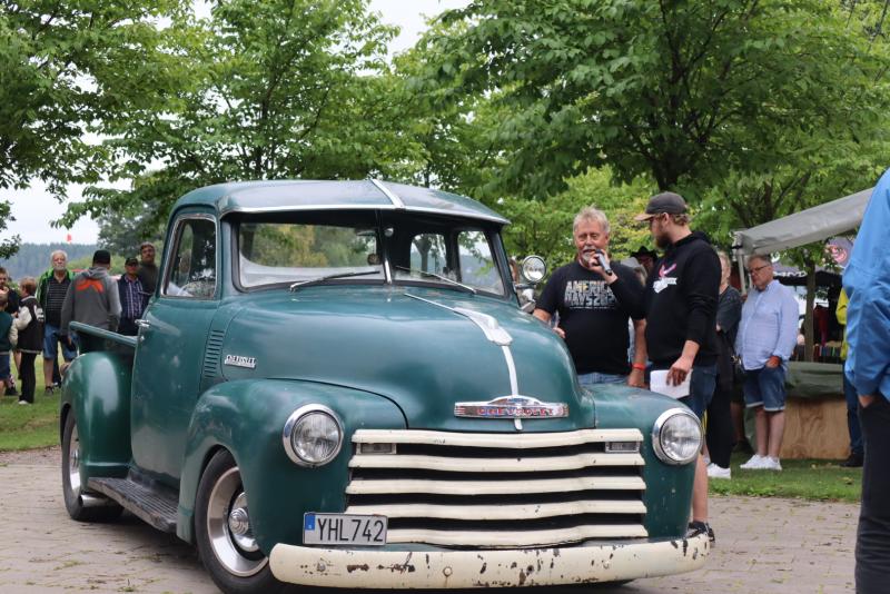 En grön Chevrolet-pickup i retrostil står parkerad utomhus, med flera personer som står i närheten och träd i bakgrunden.