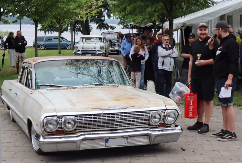 En vit veteranbil står parkerad vid ett utomhusevenemang. Människor står i närheten, varav några håller i mikrofoner och en liten pokal. I bakgrunden syns tält och träd.