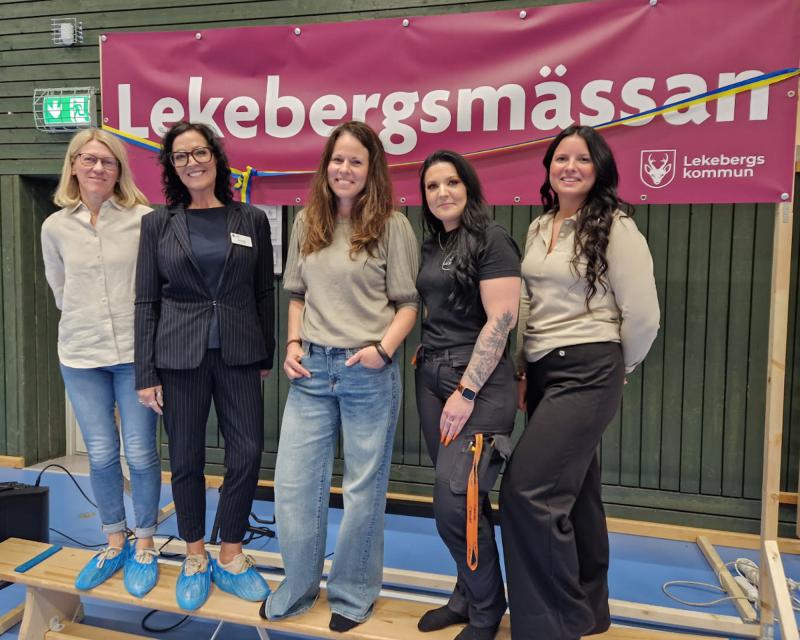 Fem kvinnor står inomhus framför en rödbrun banderoll med texten 