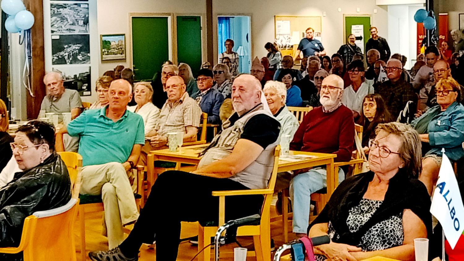 En grupp människor, de flesta äldre, sitter vid bord i en lokal och lyssnar uppmärksamt på någon som inte syns i bild.