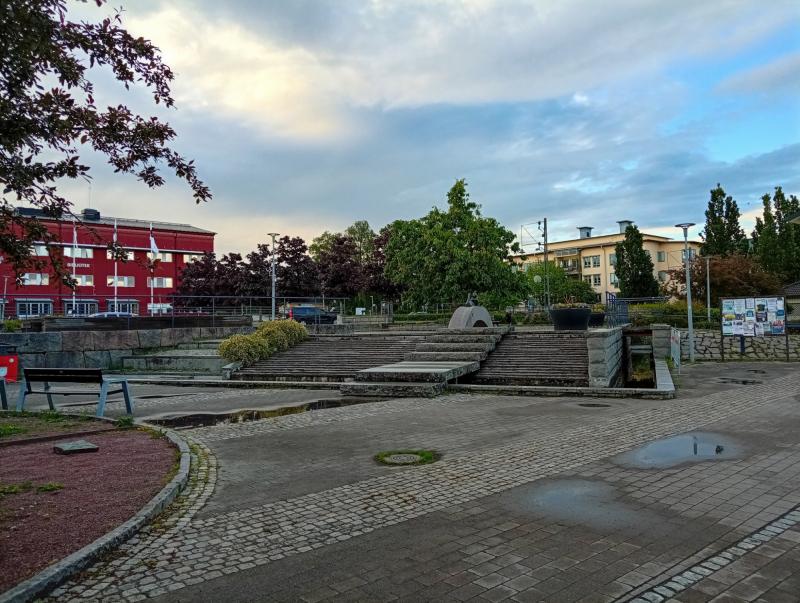 Ett stenlagt torg med trappor, träd, bänkar och närliggande byggnader under en delvis molnig himmel. En anslagstavla och stora planteringskärl syns.