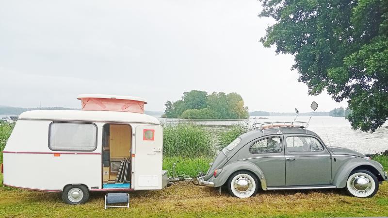 En grå veteranbil står parkerad vid en sjö och drar en liten vit husvagn med öppen dörr. I bakgrunden syns träd, gräs och vatten.