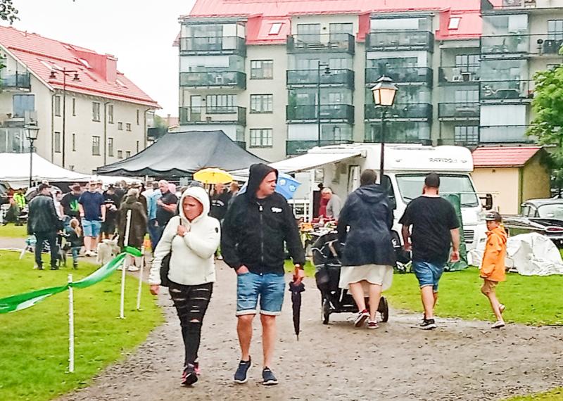 Människor går längs en stig vid ett utomhusevenemang en molnig dag, med tält, en vit skåpbil och flerbostadshus synliga i bakgrunden.
