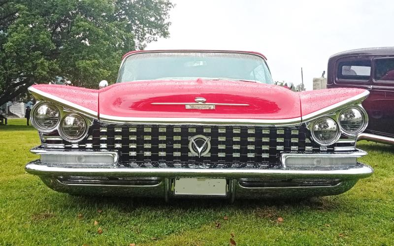 Framifrånbild av en klassisk röd Buick med kromdetaljer, parkerad på gräsmattan, där den karakteristiska kylargrillen och strålkastarna syns tydligt. I bakgrunden syns träd och en annan veteranbil.
