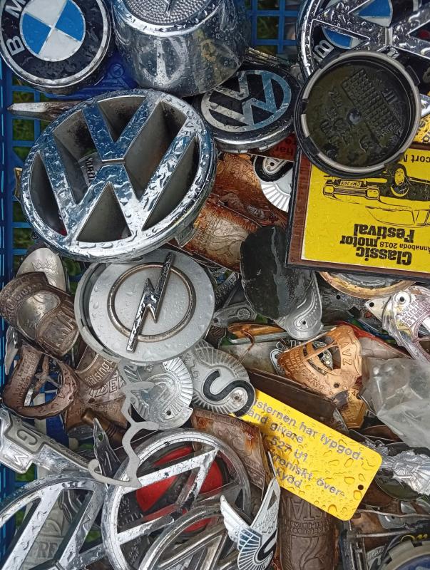 Ett urval av bilmärken och emblem, bland annat från Volkswagen, BMW och Opel, uppställda i en blå plastbricka tillsammans med små metallprydnader och ett gult kort.