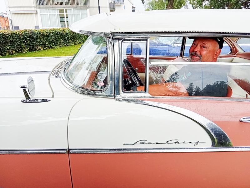 En man med hatt sitter i förarsätet i en äldre tvåfärgad Pontiac Star Chief som står parkerad nära en häck och en vit byggnad.