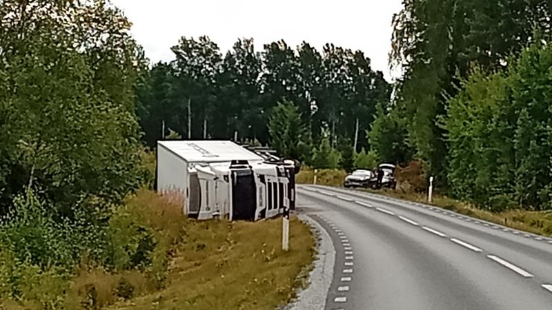 En stor vit lastbil ligger på sidan vid sidan av en kurvig landsväg, med träd och parkerade fordon i närheten.