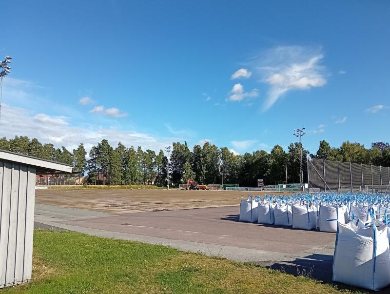 En idrottsplan utomhus som håller på att renoveras, med stora vita säckar i förgrunden, byggmaskiner i fjärran samt träd och blå himmel i bakgrunden.