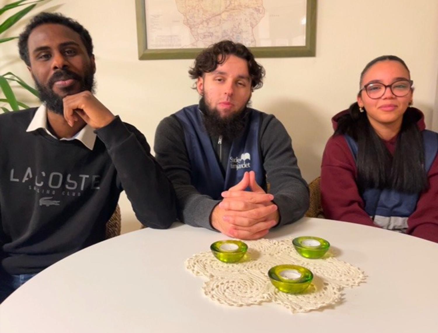 Tre personer sitter vid ett runt vitt bord med två gröna ljusstakar och virkade dukar. En inramad karta hänger på väggen bakom dem.