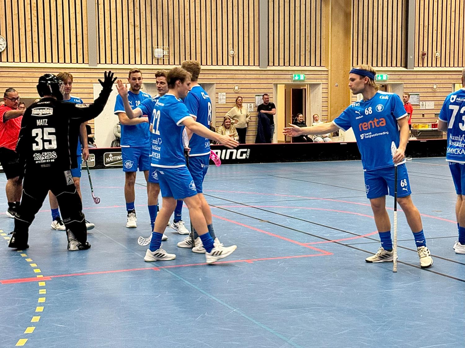 En grupp innebandyspelare i blåa uniformer och en i svart gör high five och skakar hand på en inomhusplan medan åskådare tittar på från bakgrunden.