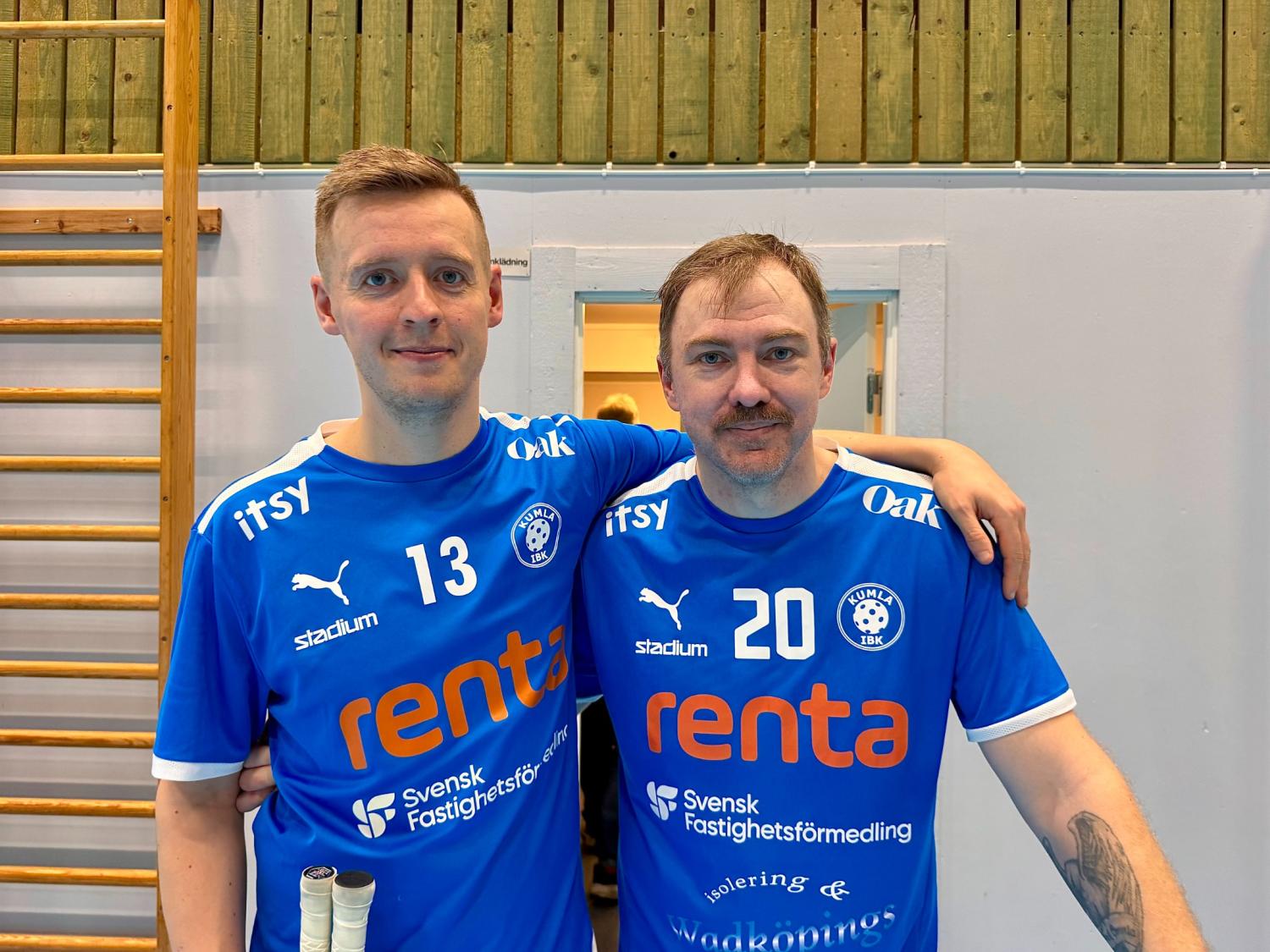 Två män i blå sporttröjor med siffrorna 13 och 20 står sida vid sida i ett inomhusgym med armarna runt varandras axlar.