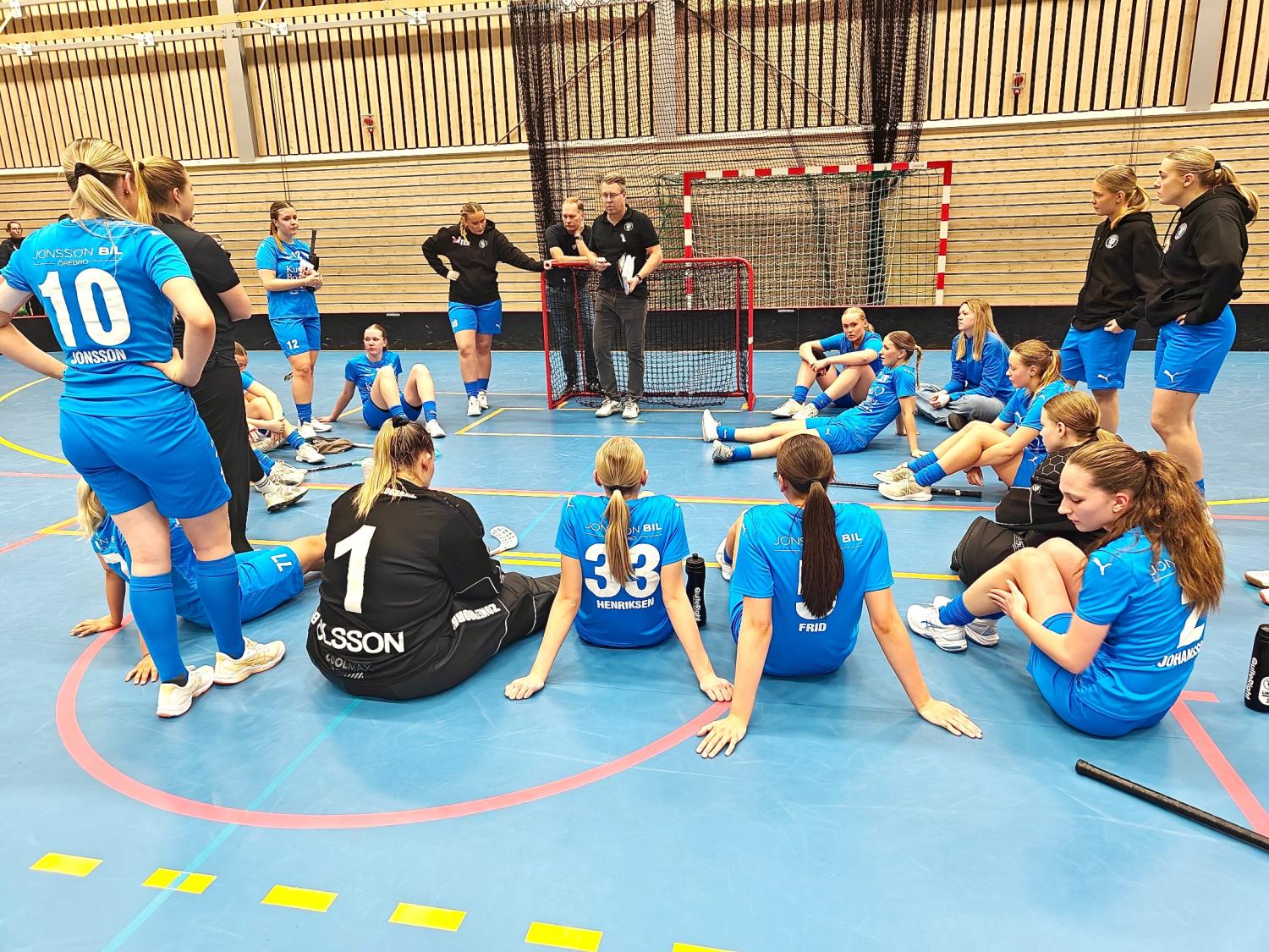 Ett damlag i innebandy i blåa dräkter sitter och lyssnar på tränarna under ett lagmöte på en inomhusplan.
