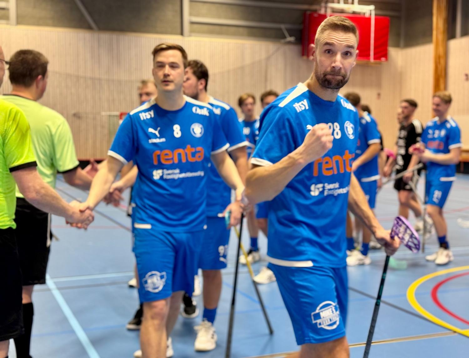 En grupp innebandyspelare i blåa dräkter skakar hand med motståndarlaget efter en match i en idrottshall.