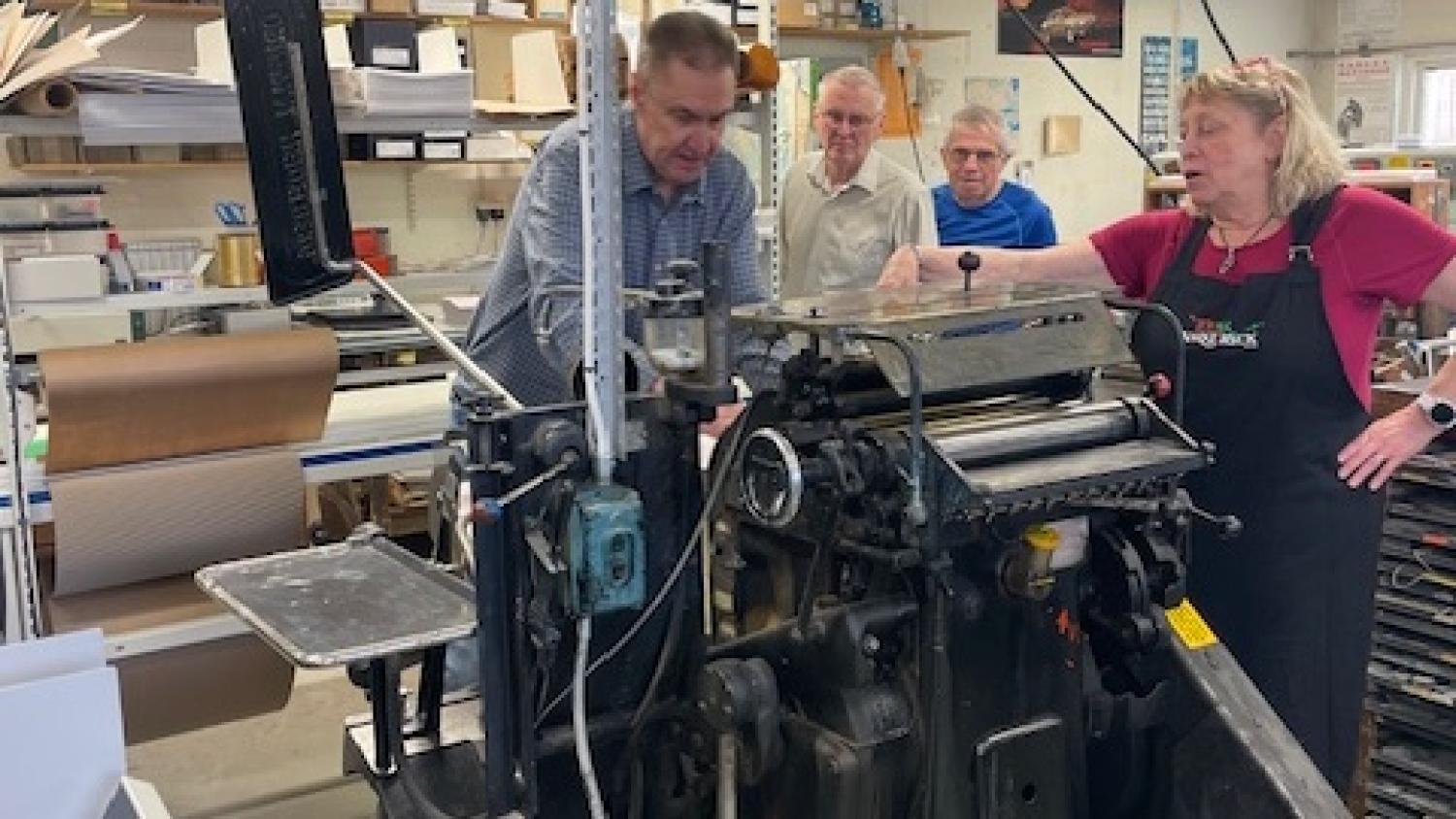 Fyra vuxna står runt en gammal tryckpress i en verkstad och tittar på när en person använder maskinen. Hyllor med förbrukningsmaterial syns i bakgrunden.