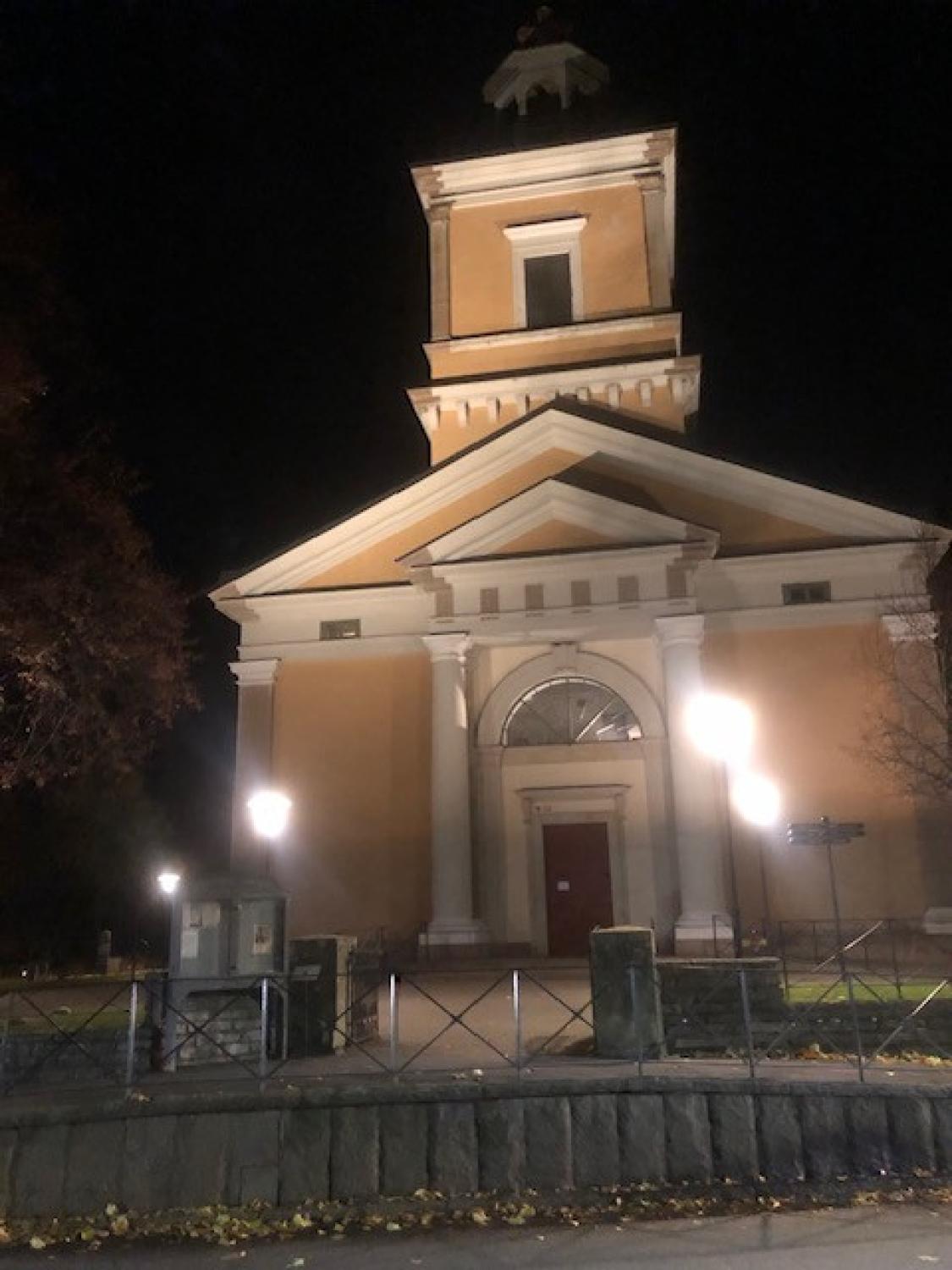 En gul kyrkobyggnad med vita pelare är upplyst på natten, med gatlyktor och nedfallna löv synliga framför.