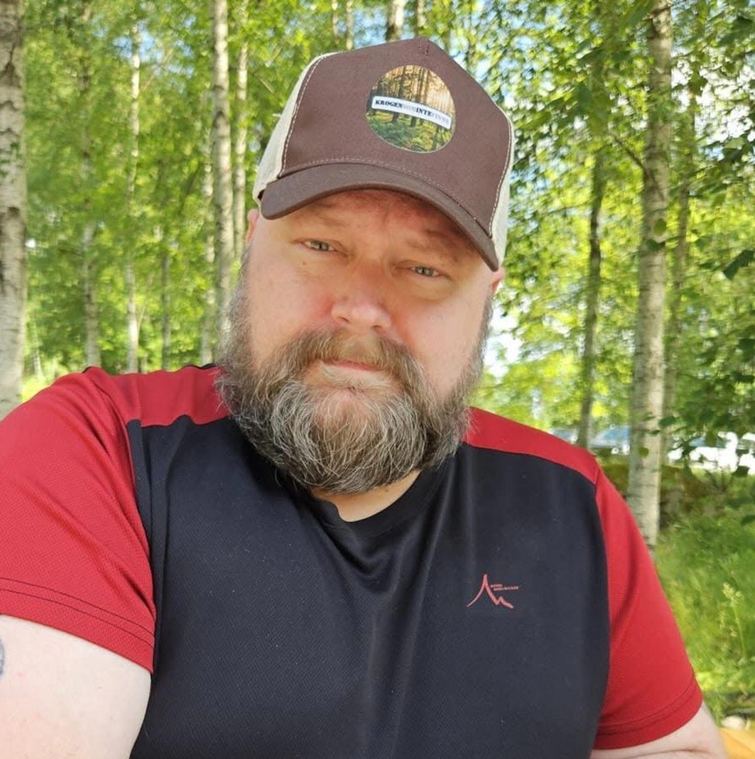 En skäggig man med brunvit keps och svartröd skjorta sitter utomhus i ett skogsparti där solljuset silar genom träden.