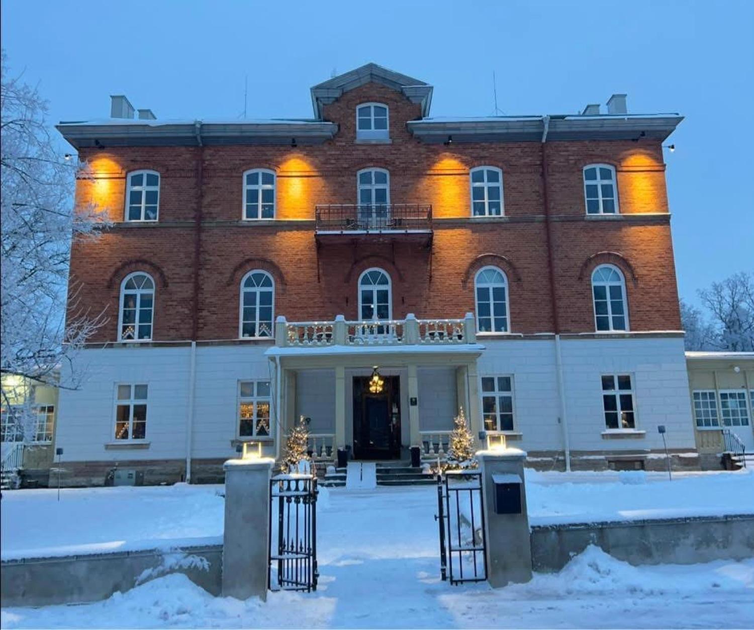 En trevåningsbyggnad i tegel och stuckatur är upplyst med utomhusbelysning, marken är täckt av snö och vid entrén står små dekorerade träd.