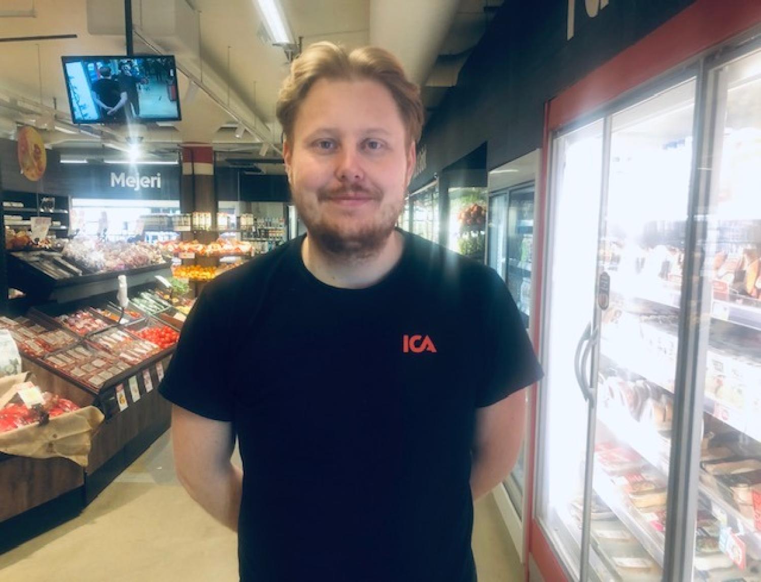 En man med en ICA-t-shirt står i en mataffär i närheten av kyl- och frysdiskar och tittar in i kameran.