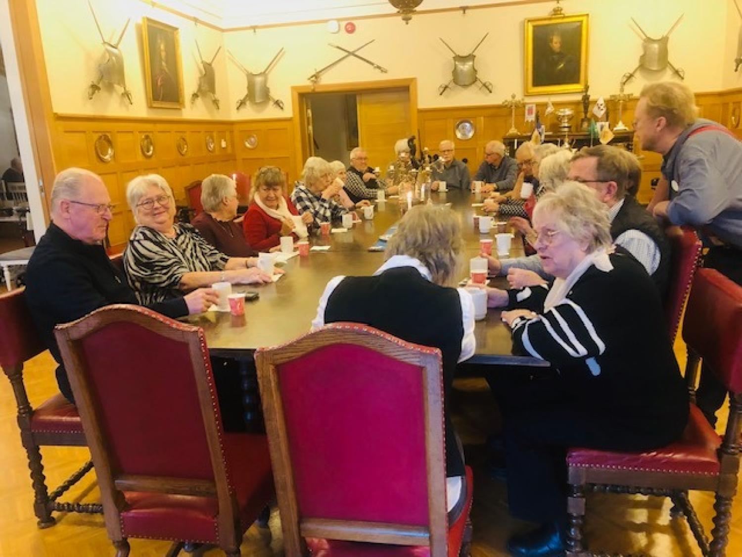 En grupp äldre människor sitter runt ett långt träbord i ett inrett rum, dricker ur koppar och samtalar.