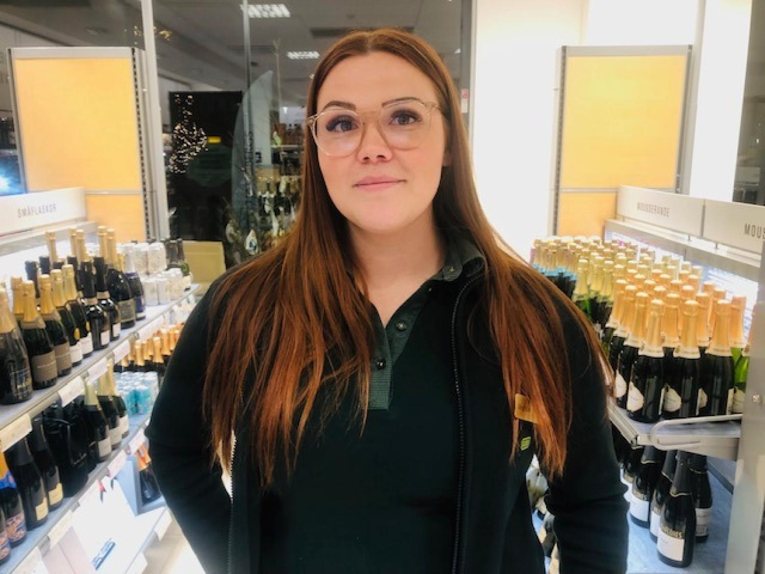 En kvinna med långt brunt hår och glasögon står i en butiksgång med flaskor med mousserande vin och champagne.
