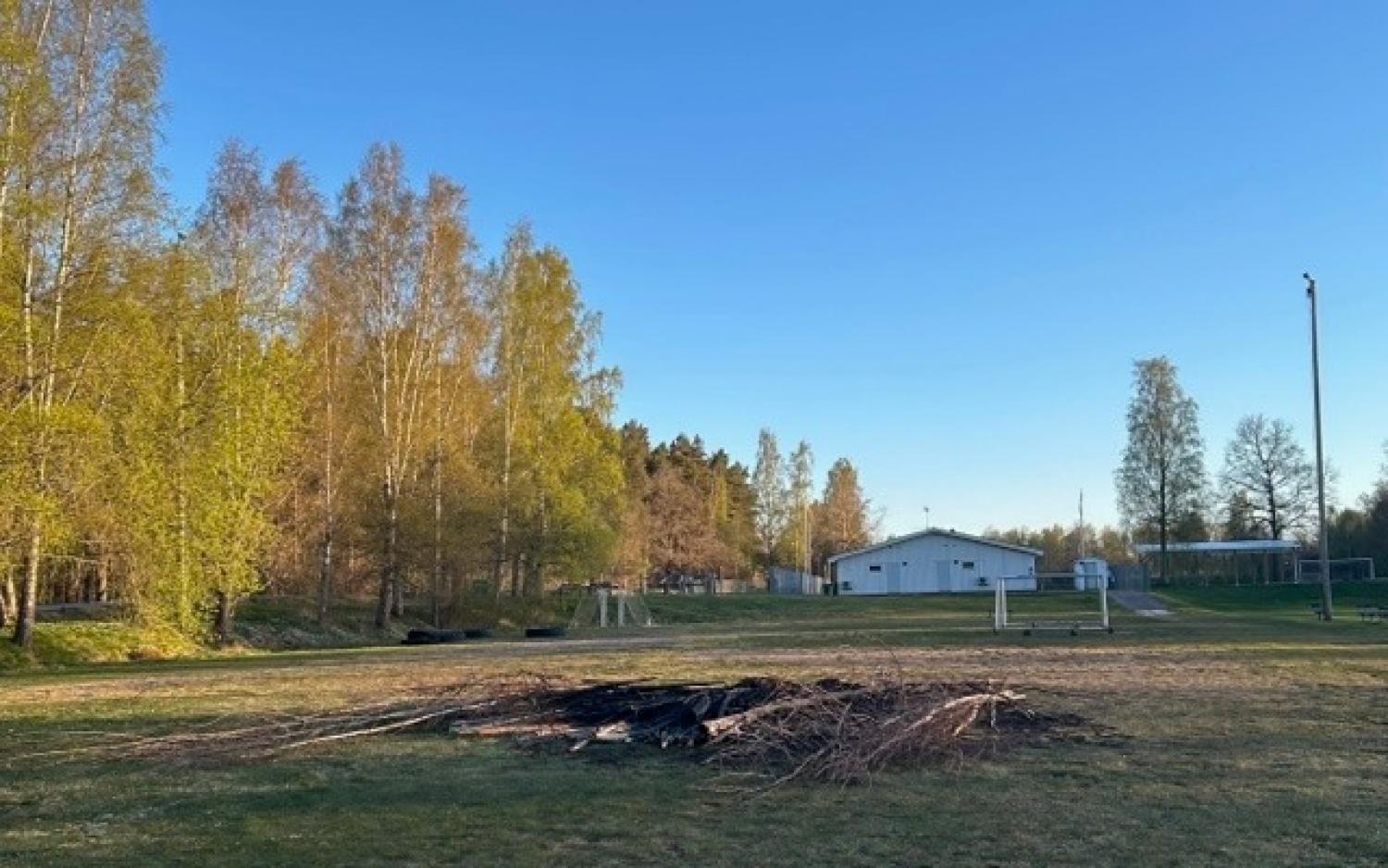 Ett gräsbevuxet fält med en hög av grenar i mitten, omgivet av träd och en vit byggnad i bakgrunden under en klarblå himmel.