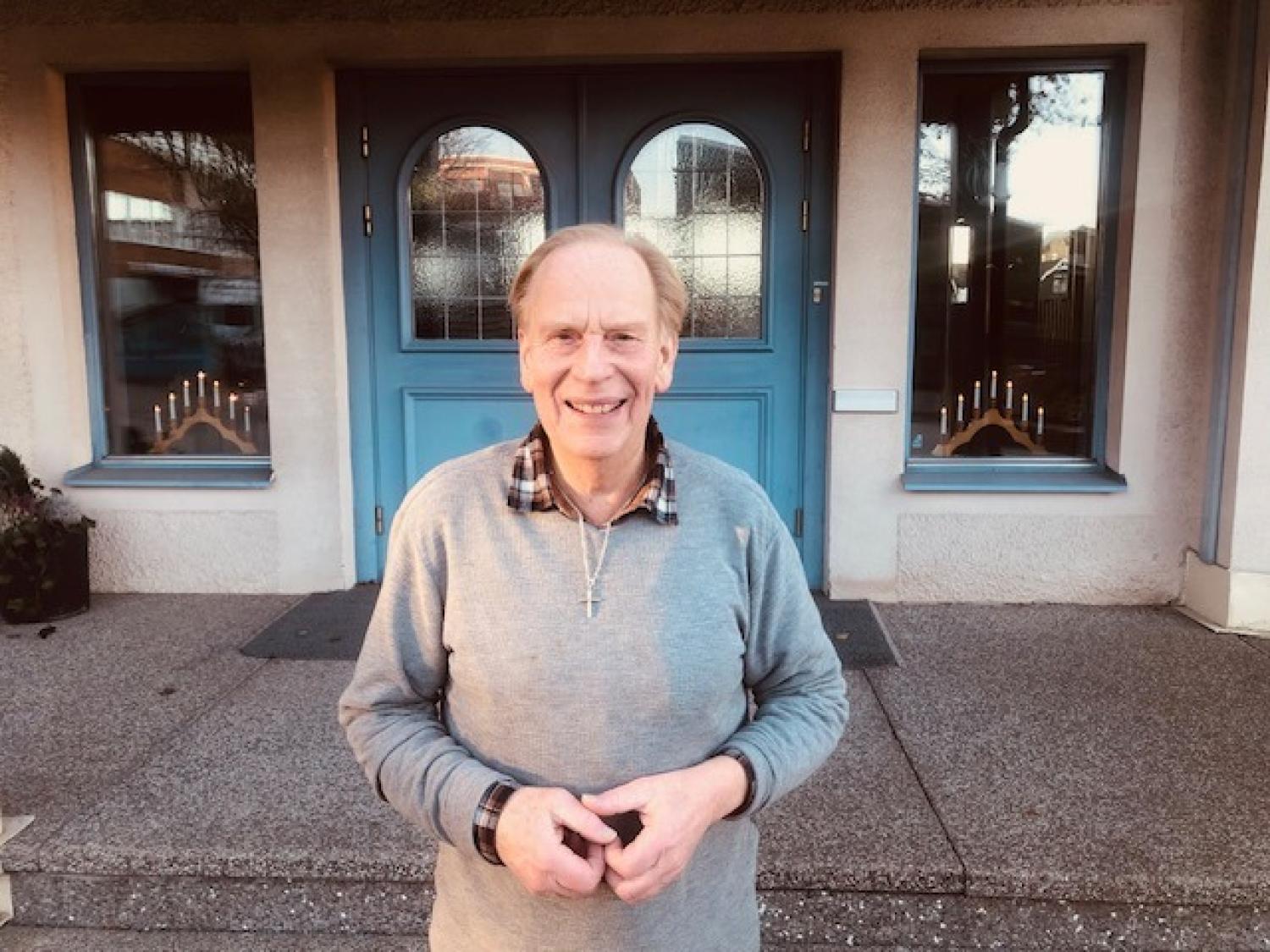En äldre man i grå tröja står leende framför blå dubbeldörrar med glaspartier och två fönster på vardera sidan.