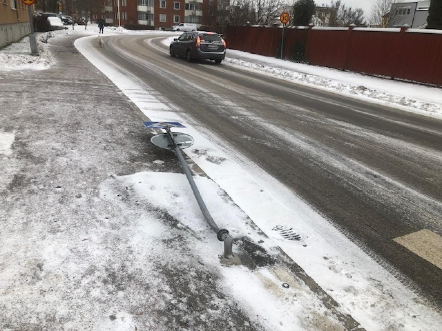 En böjd gatuskylt ligger på en snöig trottoar bredvid en krökt väg, med en bil som kör iväg och byggnader i bakgrunden.