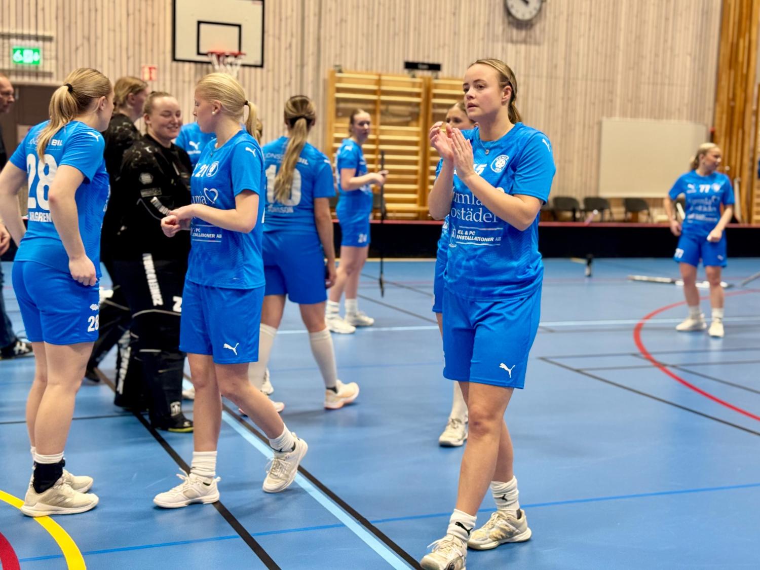 Ett damlag i inomhusidrott i blåa uniformer står och går på en idrottsplan, några spelare klappar och interagerar nära en basketkorg.