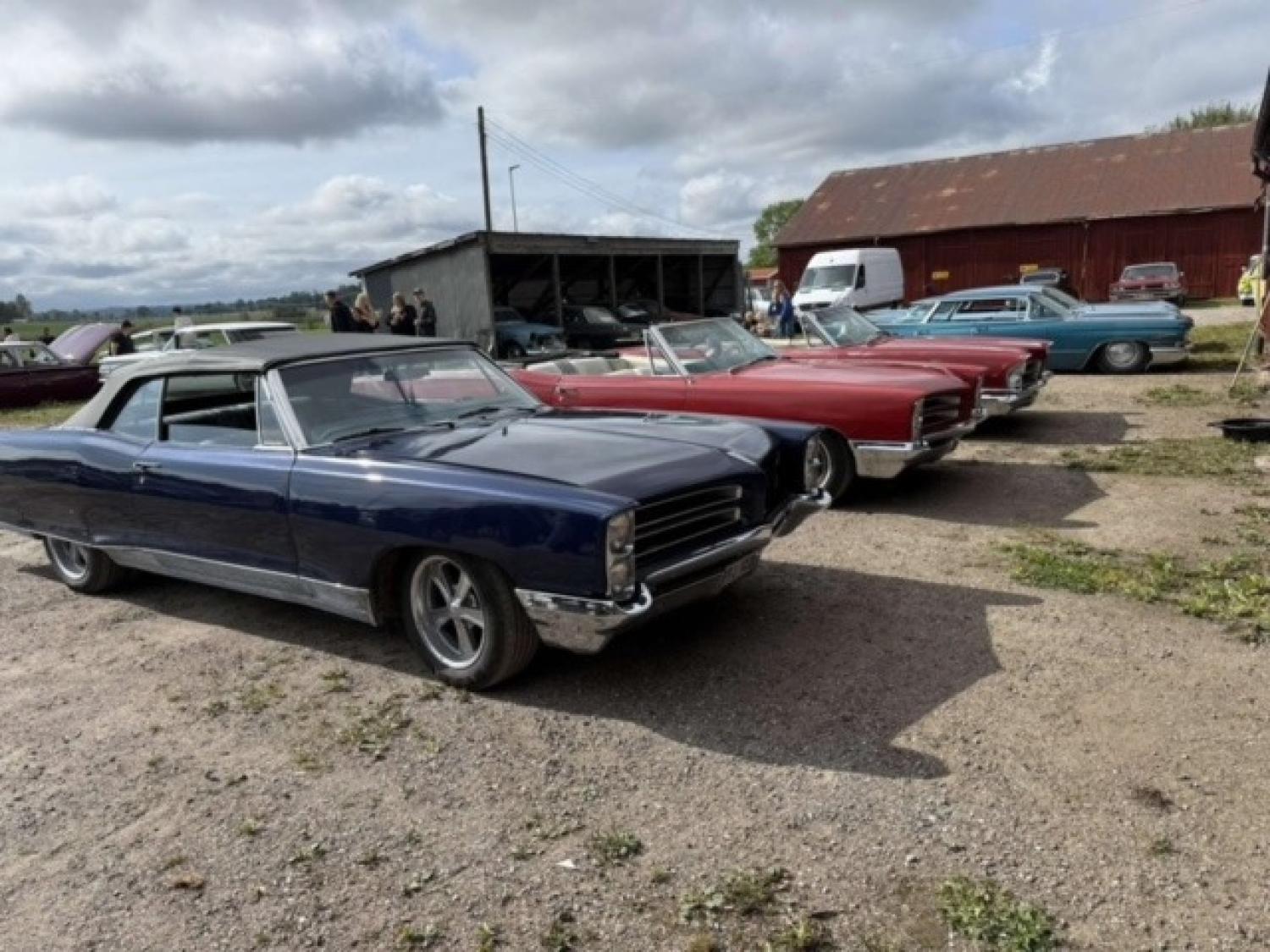 Flera klassiska cabrioletbilar i olika färger står parkerade på en parkeringsplats en molnig dag, med lador och andra fordon i bakgrunden.