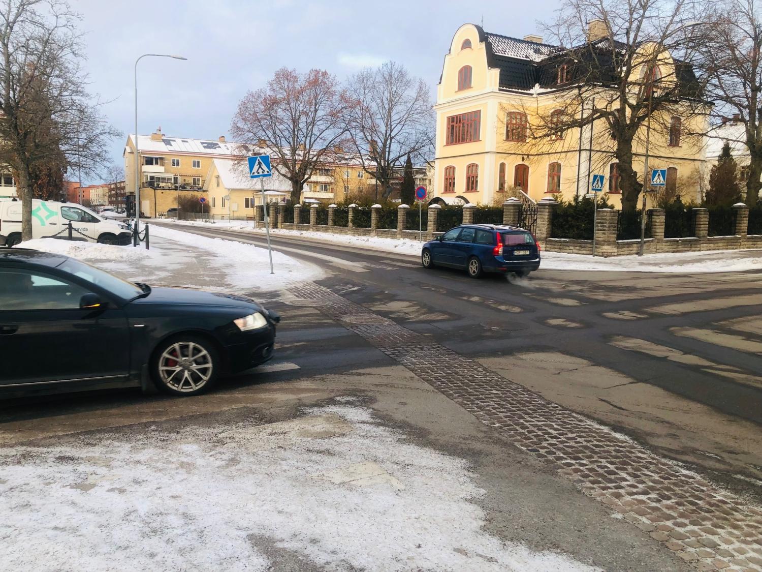 Bilar kör på en delvis snötäckt gata i ett bostadsområde med stora hus och kala träd en molnig dag.