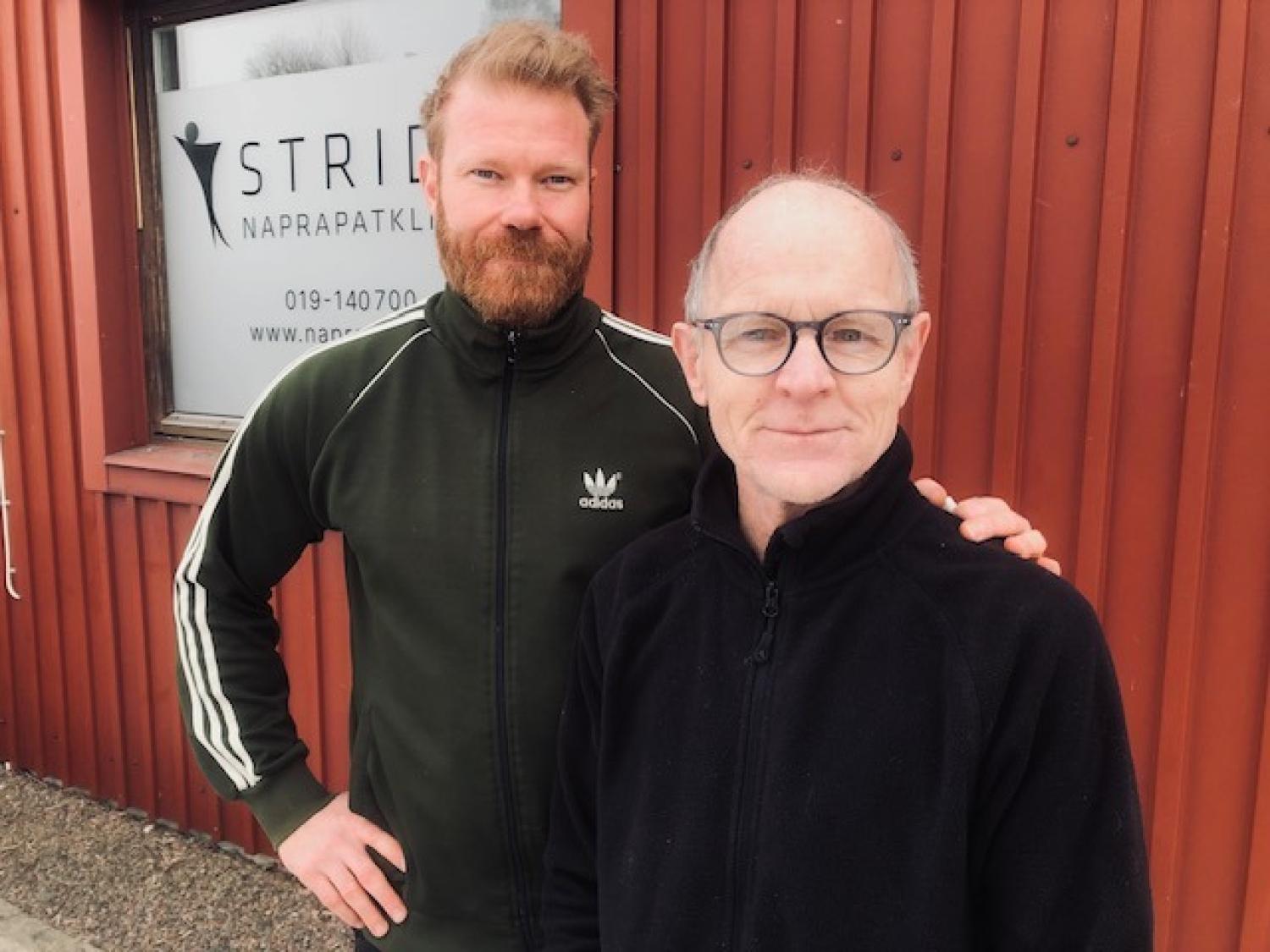 Två män står utomhus framför en röd byggnad med en klinikskylt i bakgrunden. Den ena mannen har skägg och bär en grön jacka, den andra har glasögon och en svart jacka.