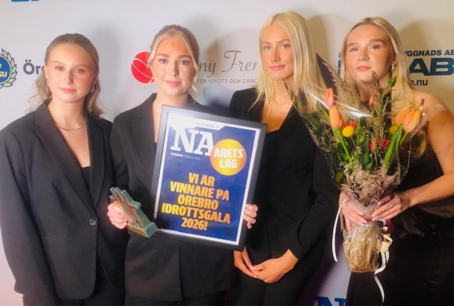 Fyra formellt klädda kvinnor står tillsammans och håller i en pokal, en blombukett och en skylt med texten 