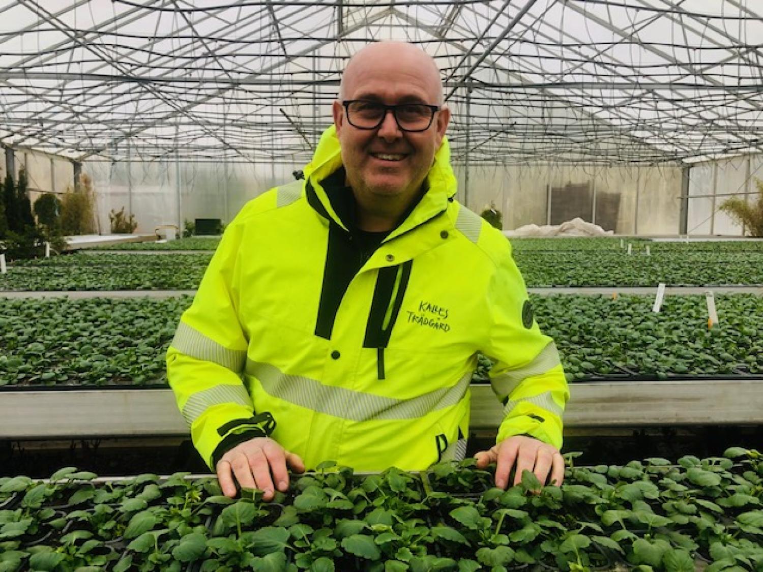 En man i en ljusgul jacka står i ett växthus och ler med händerna på rader av gröna bladväxter.