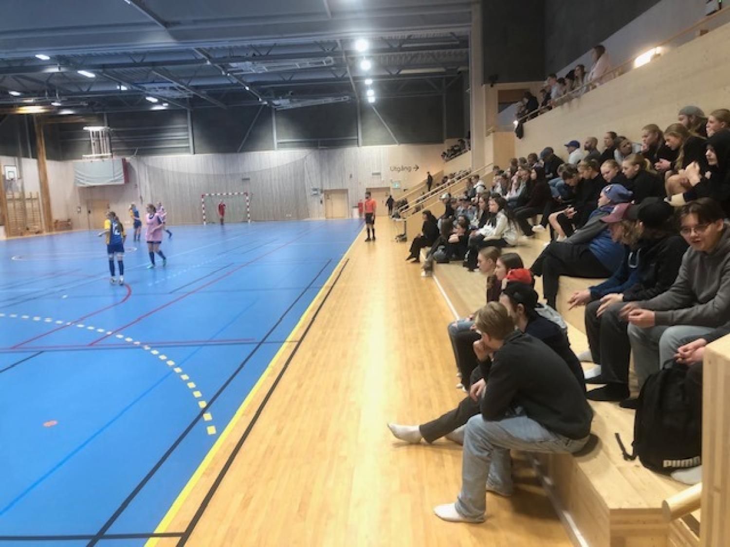 Åskådare sitter på träläktare och tittar på en handbollsmatch inomhus i en sporthall med spelare synliga på en blå plan.
