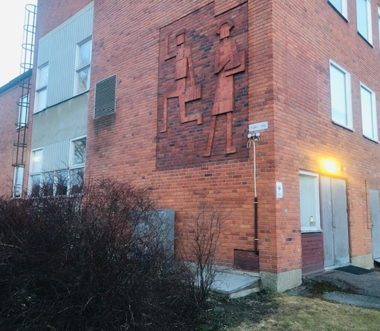 En röd tegelbyggnad med en reliefskulptur av två abstrakta människofigurer på väggen ovanför en dörröppning, bredvid några buskar och en tänd ytterbelysning.