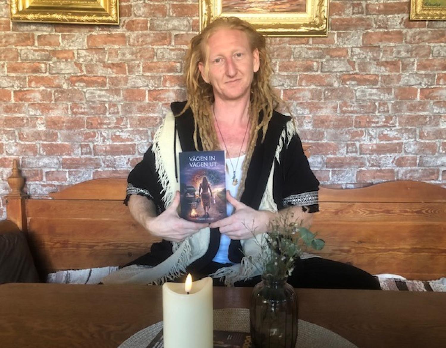 En person med långa, blonda dreadlocks sitter på en träbänk och håller i en bok. Ett tänt ljus och en liten vas står på bordet framför dem; tegelvägg och inramade bilder i bakgrunden.