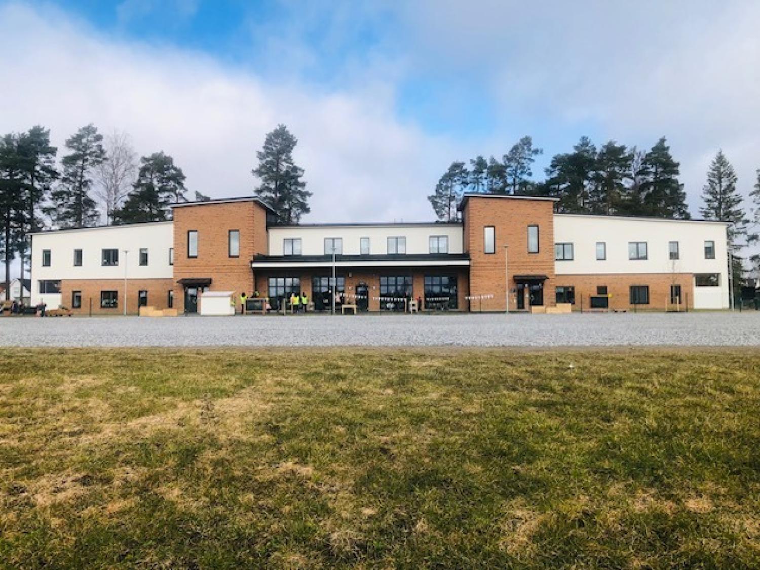 En modern tvåvåningsbyggnad med vit och brun tegelfasad, stora fönster och omgiven av träd, sedd från en gräsyta.
