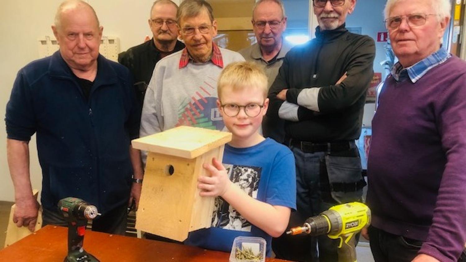 En ung pojke håller i en fågelholk av trä, omgiven av sex äldre män i en verkstad. På bordet står två borrmaskiner och en låda med skruvar.