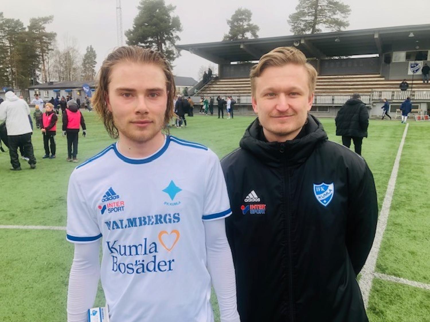 Två män står på en fotbollsplan; den ene bär en vit fotbollströja, den andre en svart jacka. Det finns människor och läktare i bakgrunden.