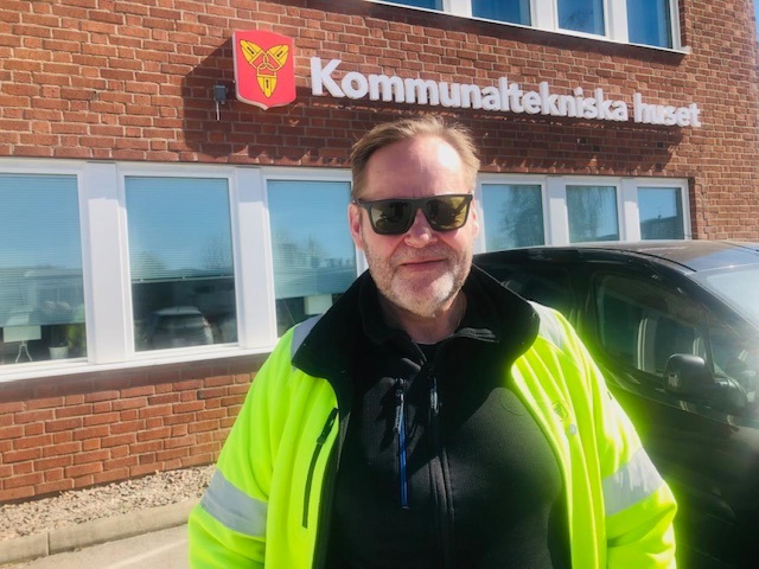 En man iförd solglasögon och varseljacka står framför en tegelbyggnad med texten 
