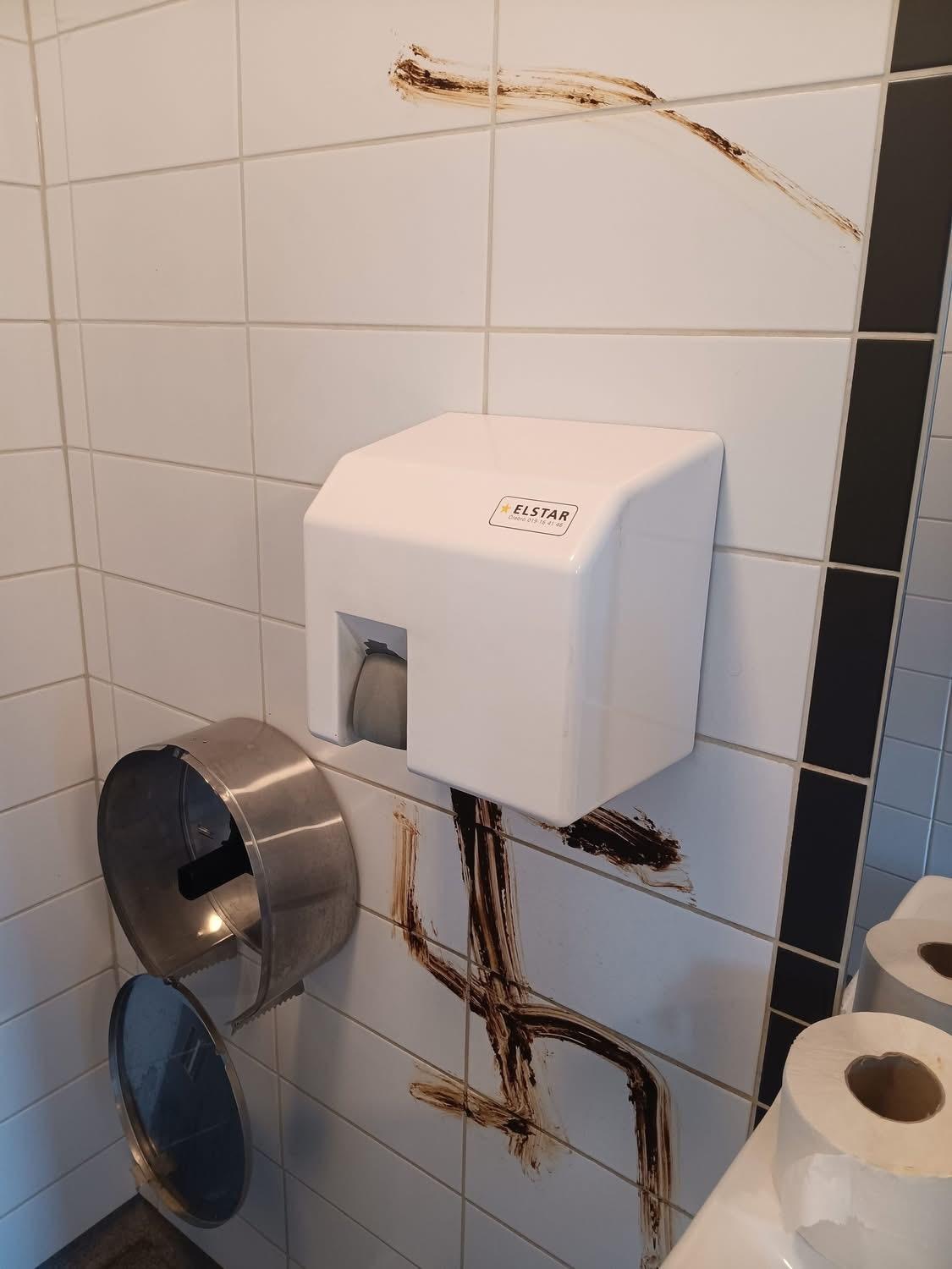 En vägg på en offentlig toalett med handtork och toalettpappersbehållare är nedsmutsad med en brun substans. Toalettpappersrullar ligger på en yta i närheten.