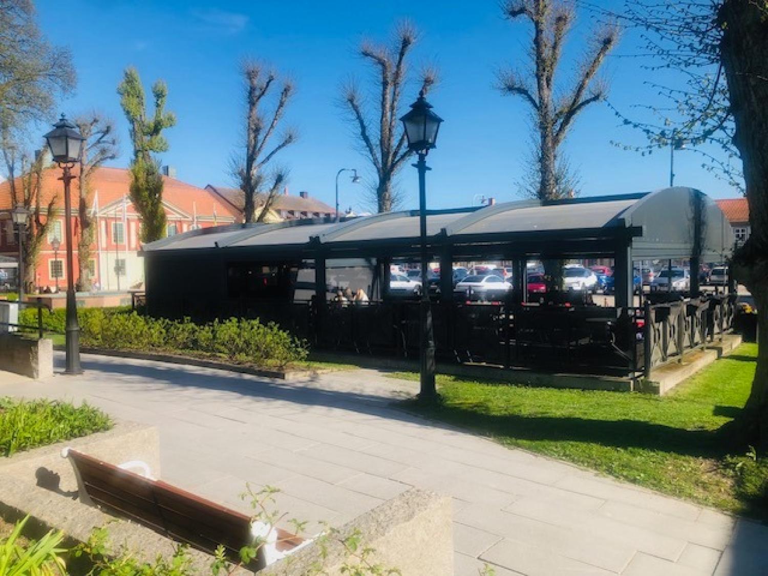Uteservering på en restaurang med svarta skärmtak, omgiven av parkerade bilar och beskurna träd, en solig dag med blå himmel.