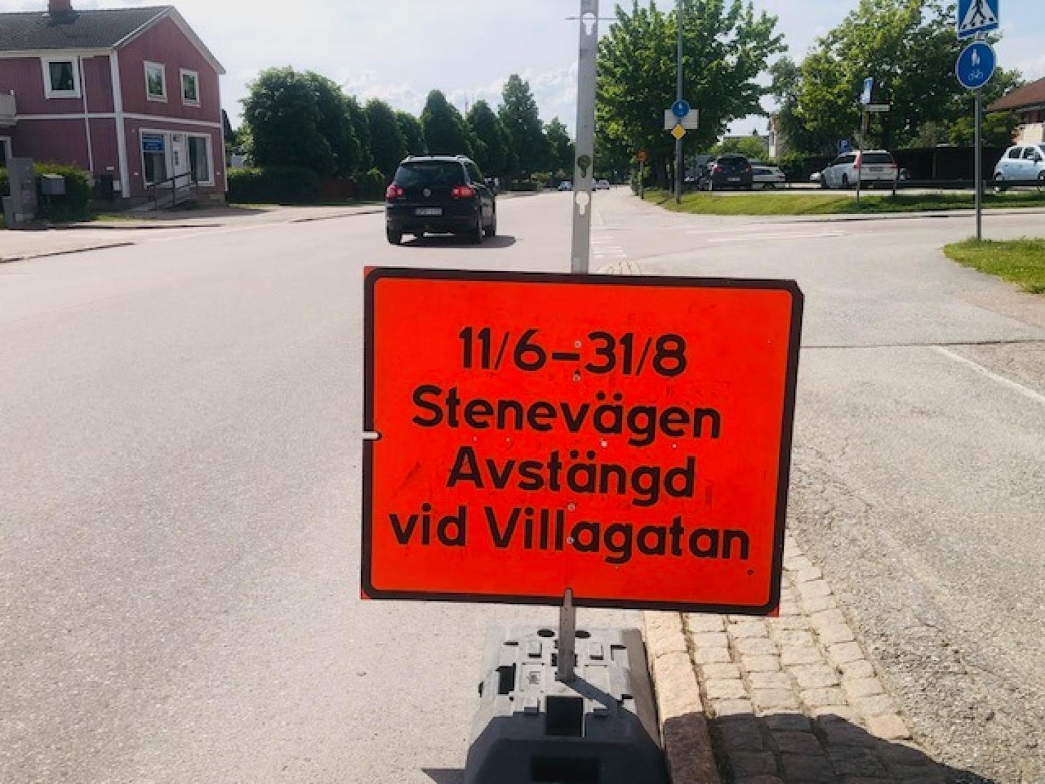 Orange vägskylt på svenska meddelar att Stenevägen är avstängd vid Villagatan från 11 juni till 31 augusti. En bil och hus syns i bakgrunden.