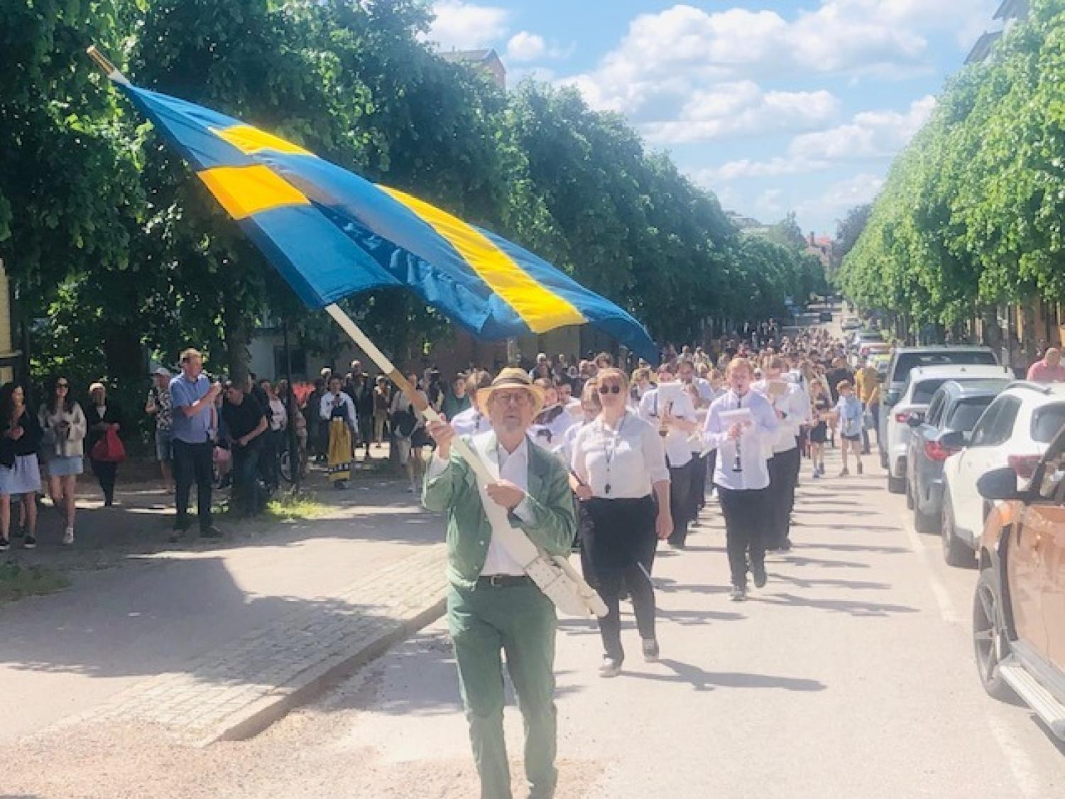 En parad marscherar längs en solig gata, ledd av en person som håller i en stor svensk flagga, med åskådare och parkerade bilar på båda sidor.
