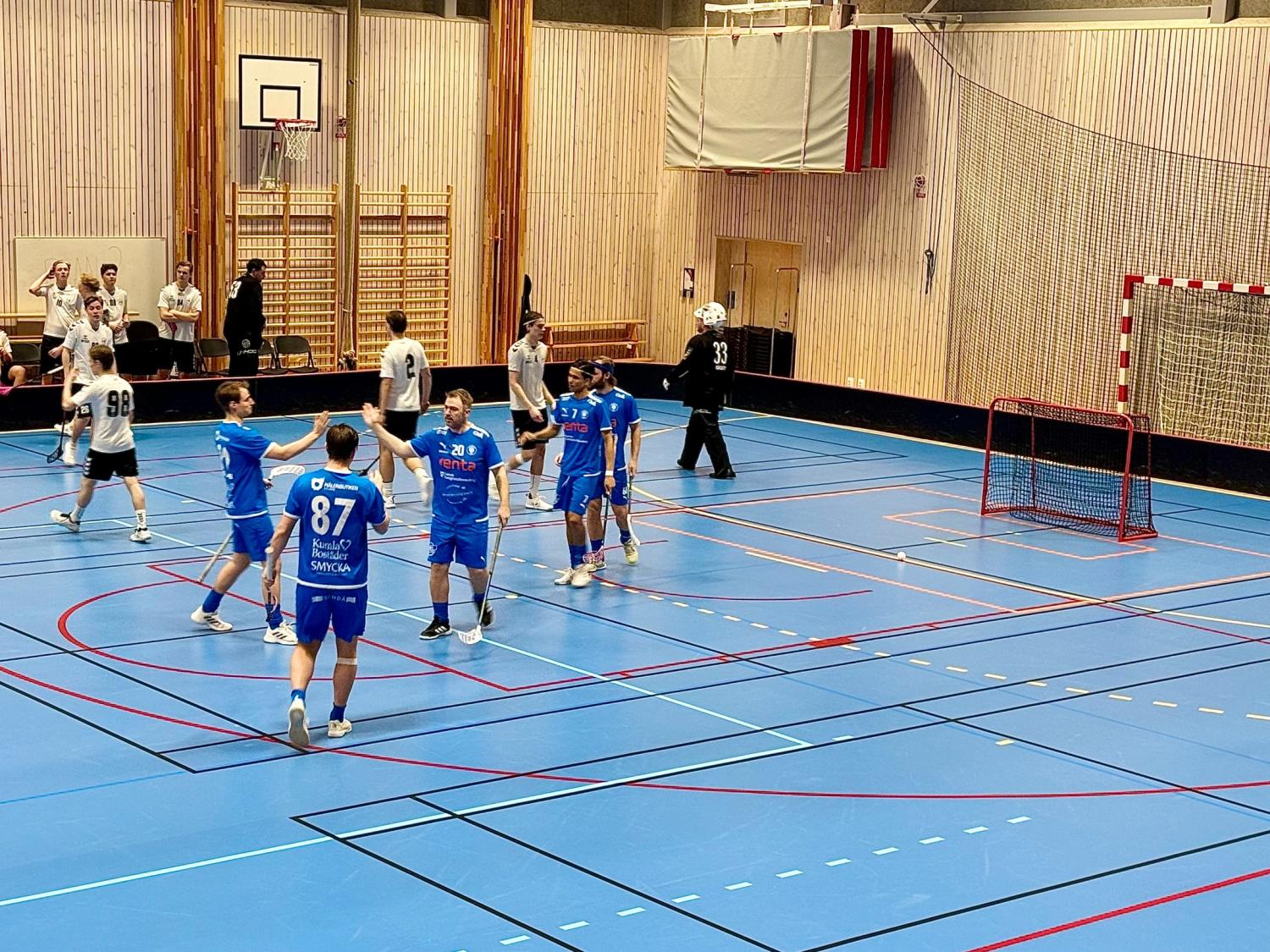 Spelare i blåvita uniformer spelar en innebandymatch i en inomhushall. Några spelare firar medan andra omgrupperar sig nära målet.
