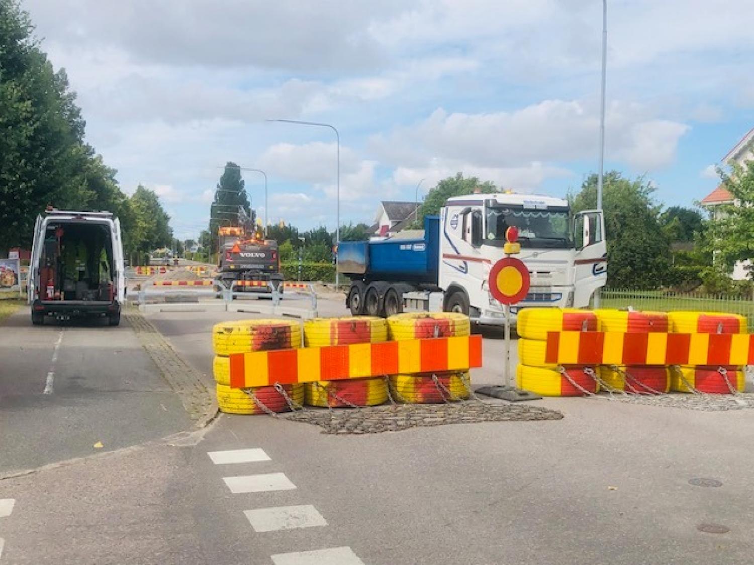 En väg under byggnation med avspärrningar som hindrar tillträde, lastbilar parkerade i närheten och maskinutrustning synlig i bakgrunden en molnig dag.