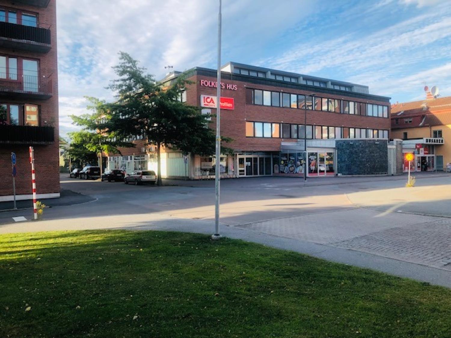 En modern trevåningsbyggnad i tegel vid ett gatuhörn rymmer en ICA-butik och andra affärer, med parkerade bilar och en gräsplätt i förgrunden.